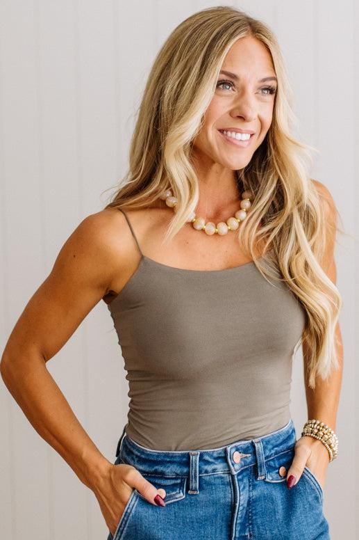 A smiling blonde woman wearing a fitted taupe-brown spaghetti-strap camisole top and blue jeans, standing against a light neutral paneled wall.
