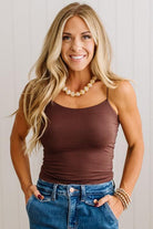 A smiling blonde woman wearing a fitted brown camisole tank top and blue jeans, standing against a light paneled wall.
