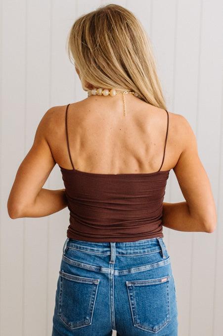 A woman shown from behind wearing a brown ruched spaghetti-strap tank top and blue jeans, standing against a white paneled wall.