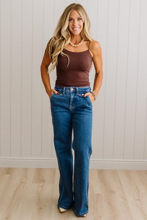 A woman modeling a brown ribbed camisole tank top and high-waisted, wide-leg blue jeans, standing against a white paneled wall on a light wood floor.