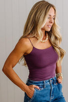 A woman wearing a fitted purple spaghetti-strap camisole top and blue jeans, standing against a light gray paneled wall.