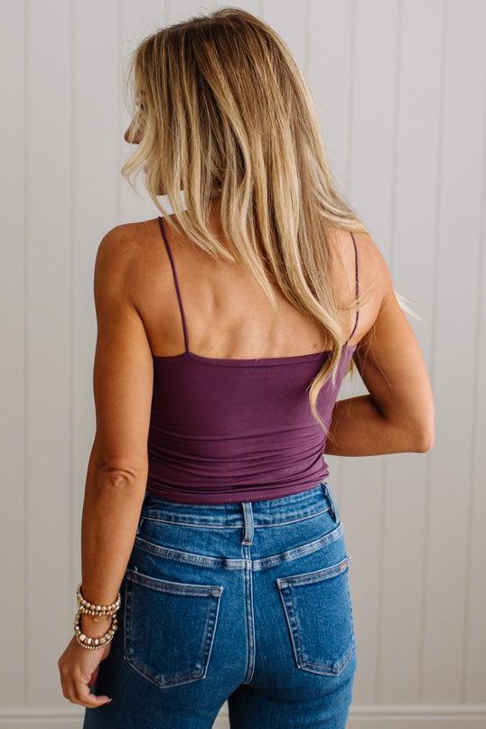 A woman shown from behind wearing a purple spaghetti-strap camisole top and blue jeans, standing against a white paneled wall.