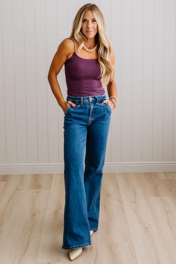 A woman modeling a pair of high-waisted, wide-leg blue jeans with a purple camisole top, standing in a room with light wood floors and white paneled walls.
