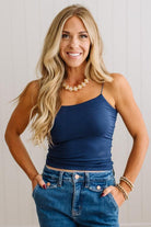 A smiling blonde woman wearing a navy blue spaghetti-strap camisole top and blue jeans, standing against a light paneled wall.