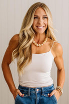 A smiling blonde woman wearing a white spaghetti-strap camisole tank top and blue jeans, accessorized with a pearl necklace and stacked bracelets, posing with hands in her pockets against a light neutral background.