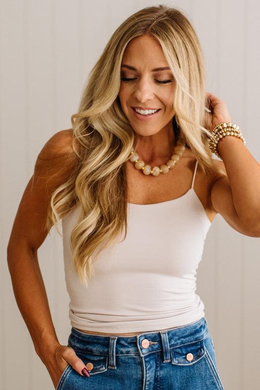 A woman with long blonde wavy hair wearing a fitted off-white spaghetti-strap camisole top and blue jeans, accessorized with a pearl necklace and gold bracelets, posing against a light beige paneled wall.