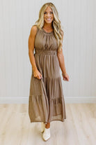 A woman modeling a sleeveless, tiered midi dress in a brown and beige gingham pattern, standing in front of a white paneled wall and light wood floor.