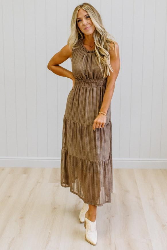 A woman modeling a sleeveless brown tiered midi dress with a subtle gingham pattern, paired with white ankle boots, standing in front of a white paneled wall and light wood floor.