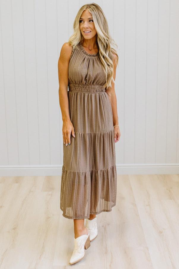 A woman modeling a sleeveless tiered midi dress in a brown and beige gingham pattern, paired with white cowboy boots, standing in a room with light wood floors and white paneled walls.
