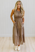 A woman modeling a sleeveless, tiered brown gingham maxi dress with side pockets, paired with white pointed-toe boots, standing against a white paneled wall and light wood floor.