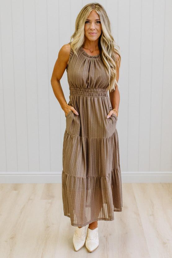 A woman modeling a sleeveless, tiered brown gingham maxi dress with side pockets, paired with white pointed-toe boots, standing against a white paneled wall and light wood floor.