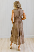 A woman modeling a sleeveless, tiered brown gingham maxi dress with side pockets, paired with white pointed-toe boots, standing against a white paneled wall and light wood floor.