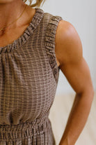 A woman modeling a sleeveless, tiered brown gingham maxi dress with side pockets, paired with white pointed-toe boots, standing against a white paneled wall and light wood floor.