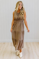 A woman modeling a sleeveless, tiered midi dress in a brown and beige gingham pattern, paired with white ankle boots, standing in front of a white paneled wall and light wood floor.