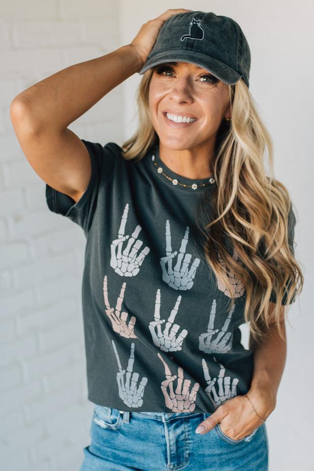 Woman wearing a black t-shirt with hand gesture patterns and a matching cap against a white brick wall.