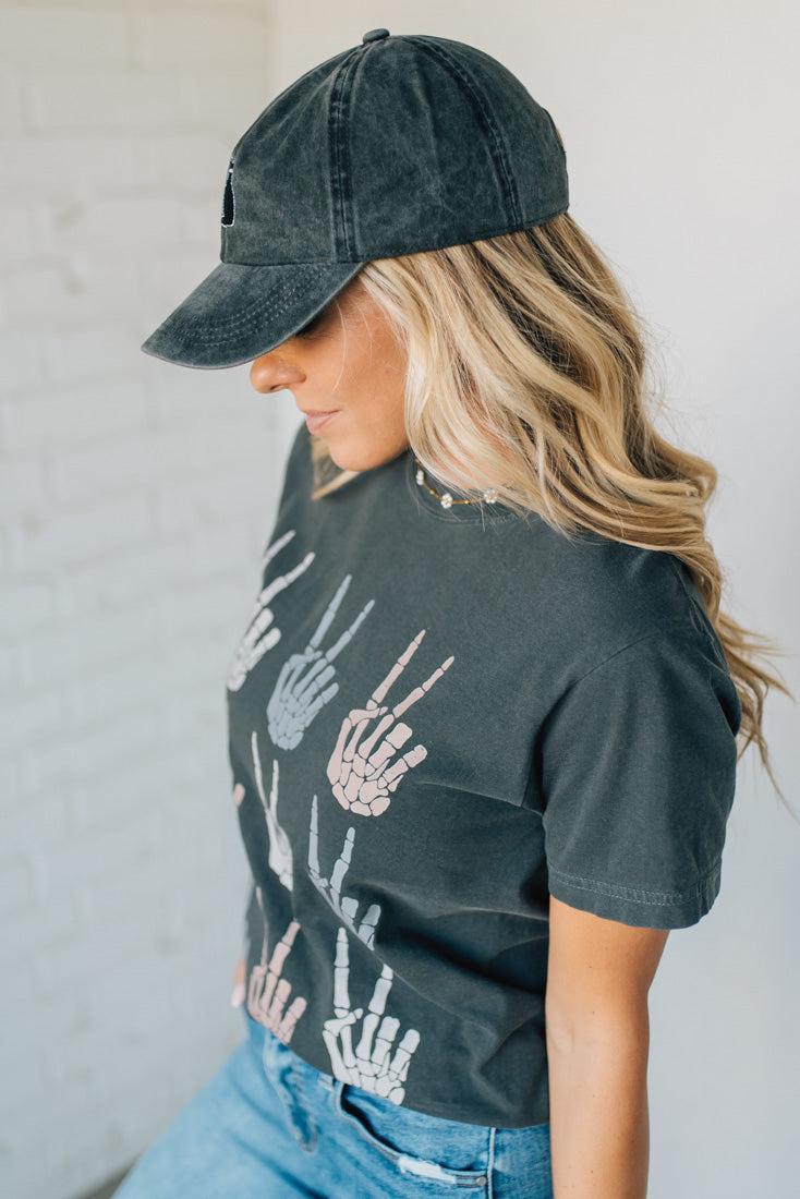 Woman wearing a dark gray t-shirt with skeleton hand designs and a matching cap.
