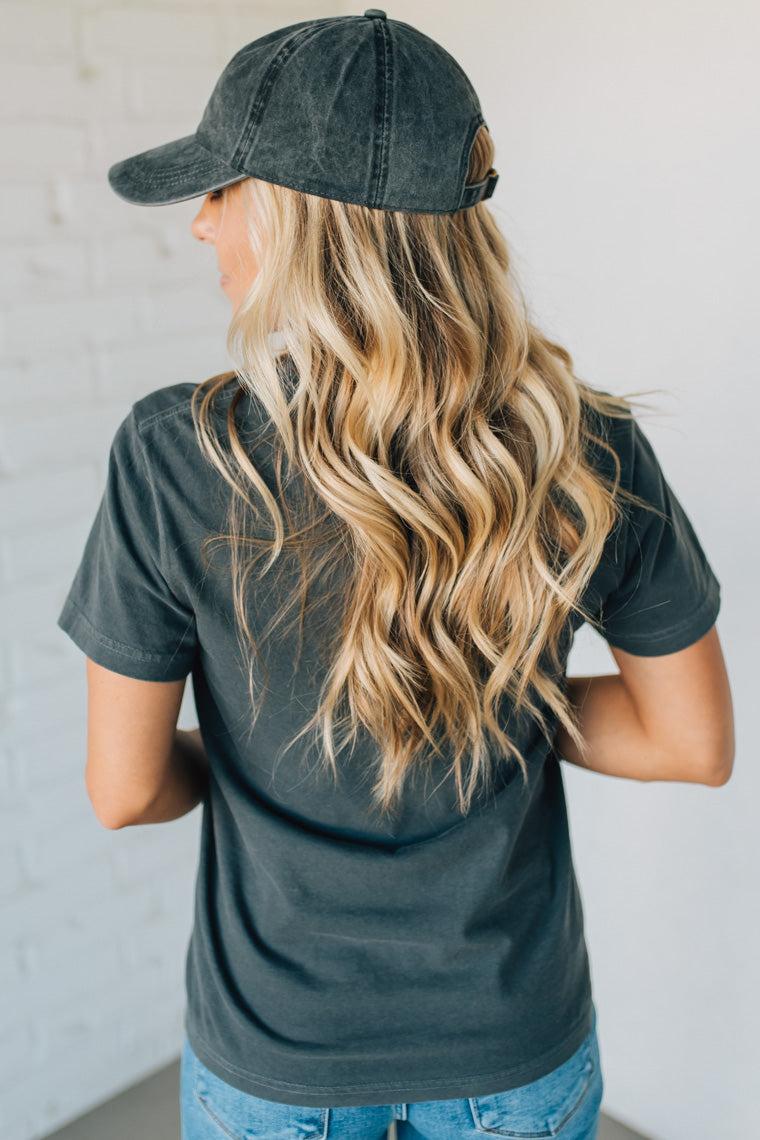Person wearing a dark green t-shirt and cap with long blonde hair, standing against a light gray wall.