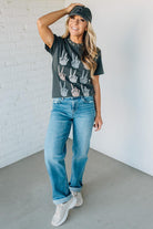 Woman wearing a graphic t-shirt, jeans, and a cap against a white brick wall.