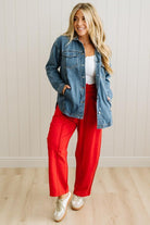 Woman wearing a denim jacket and red pants standing against a white paneled wall.
