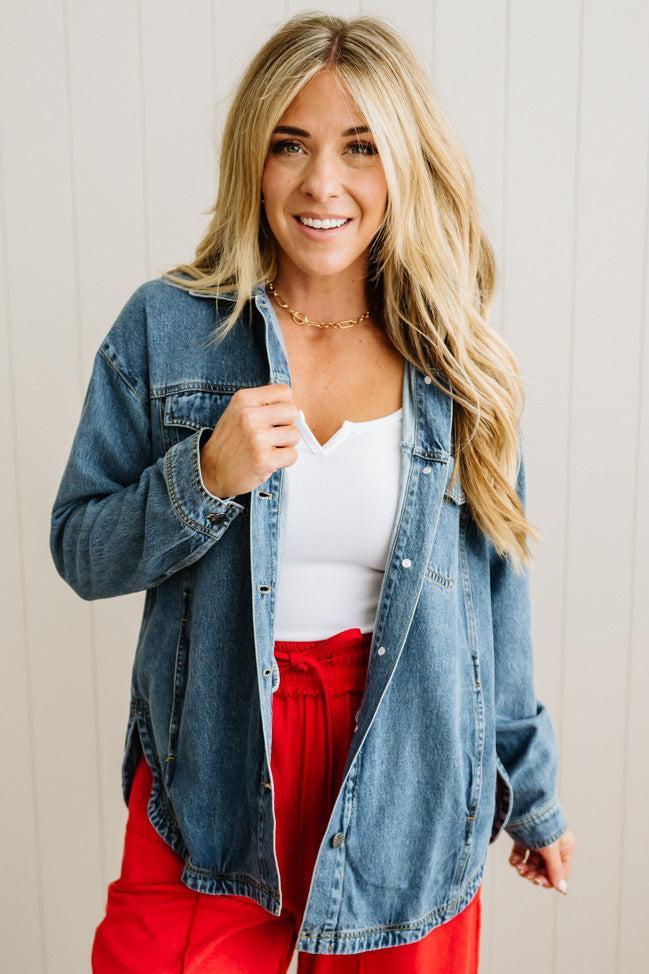 Woman wearing a denim jacket, white top, and red pants against a light wooden panel background