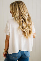 Person wearing a sand beige t-shirt and blue jeans with blonde hair, standing against a neutral background.