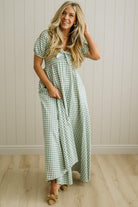 Woman wearing a green checkered dress standing against a light wooden paneled wall.