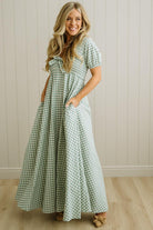 Woman wearing a green and white checkered dress standing against a light wooden paneled wall.
