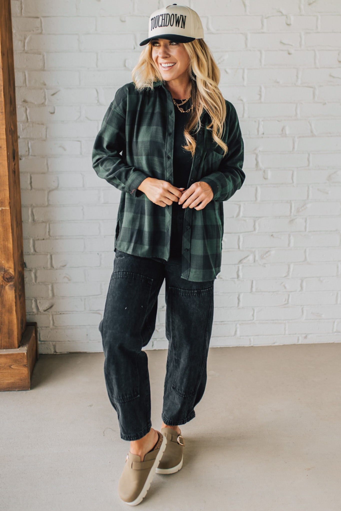 Woman wearing a green plaid shirt, black pants, and a cap indoors against a white brick wall.