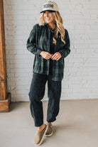 Woman wearing a green plaid shirt, black pants, and a cap indoors against a white brick wall.