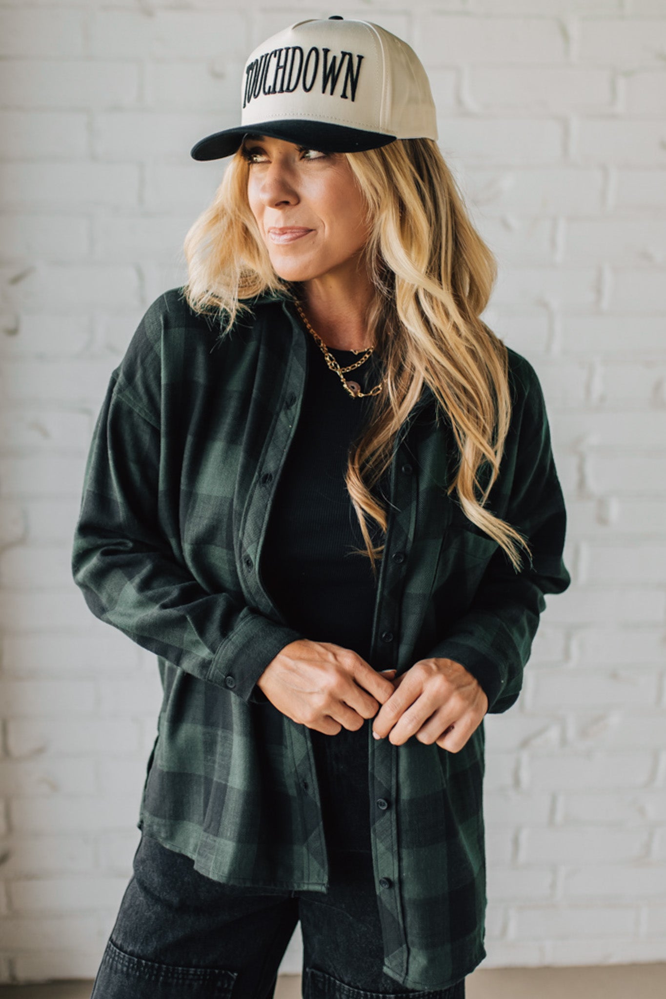 Woman wearing a green plaid shirt, black pants, and a cap with 'Touchdown' text against a white brick wall.