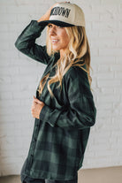 Woman wearing a green plaid shirt and beige cap with text against a white brick wall.