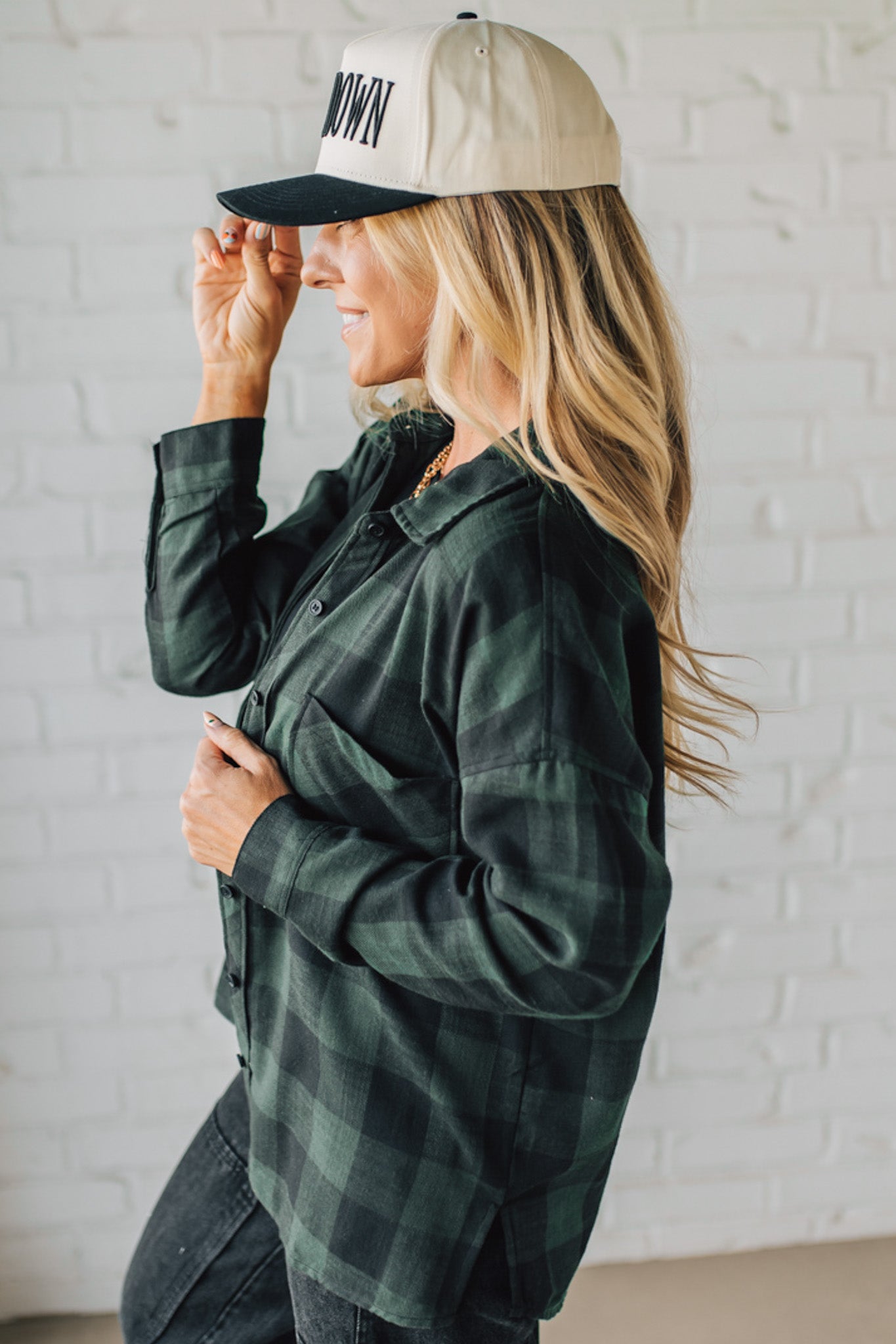 Woman wearing a green plaid shirt and beige cap with text, standing against a white brick wall.