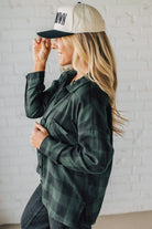 Woman wearing a green plaid shirt and beige cap with text, standing against a white brick wall.