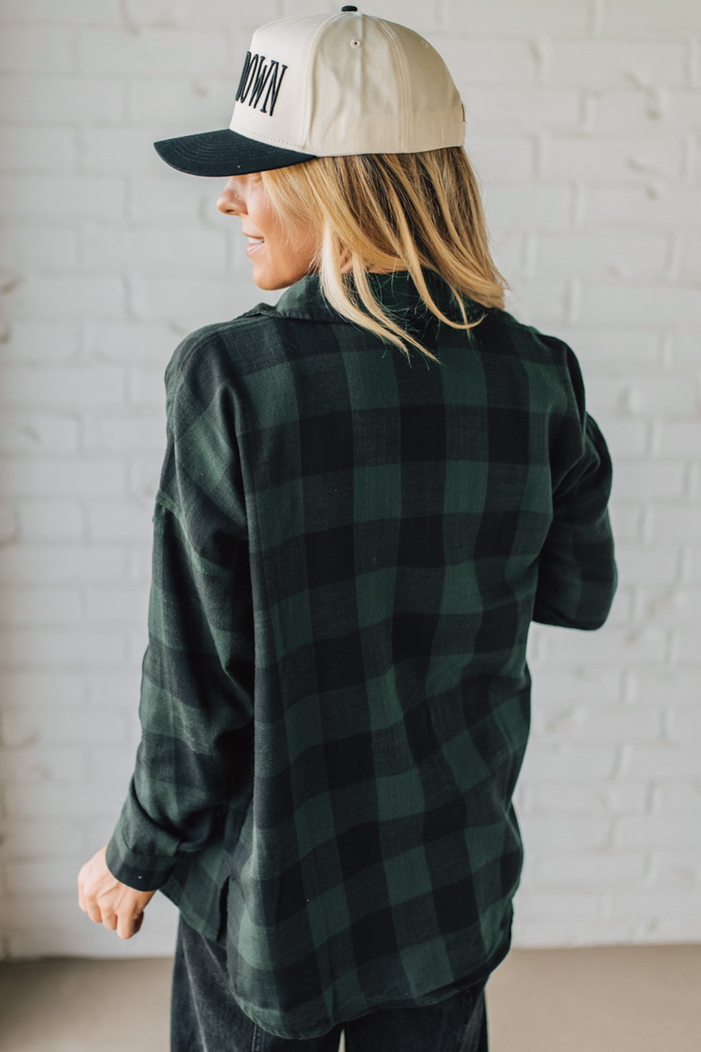 Person wearing a green plaid shirt and cap with 'OWN' text against a white brick wall.