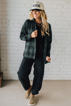 Woman wearing a plaid shirt, black pants, and a cap with a white brick wall background