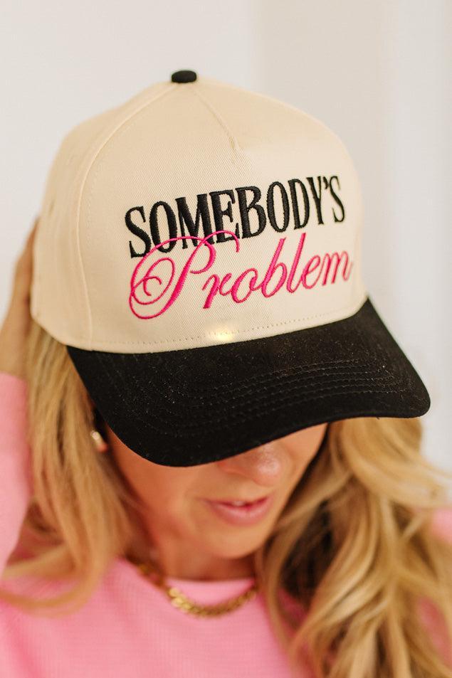Blonde woman wearing a cream and black trucker hat with "SOMEBODY'S PROBLEM" embroidered at the front in pink and black.