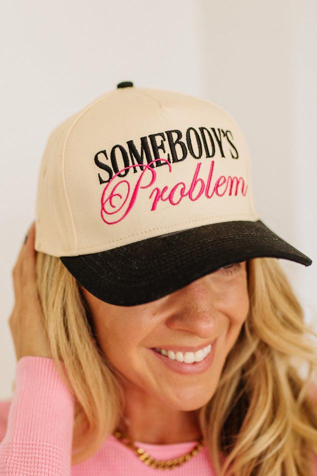 Blonde woman wearing a cream and black trucker hat with "SOMEBODY'S PROBLEM" embroidered at the front in pink and black.
