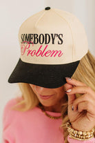 Blonde woman wearing a cream and black trucker hat with "SOMEBODY'S PROBLEM" embroidered at the front in pink and black.