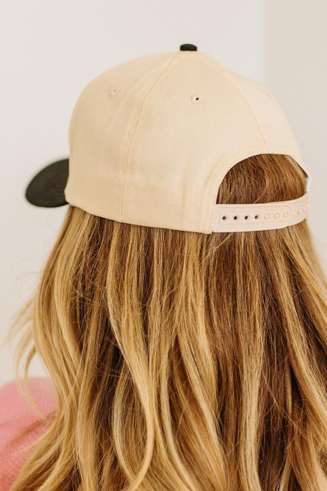 Blonde woman wearing a cream and black trucker hat with "SOMEBODY'S PROBLEM" embroidered at the front in pink and black.