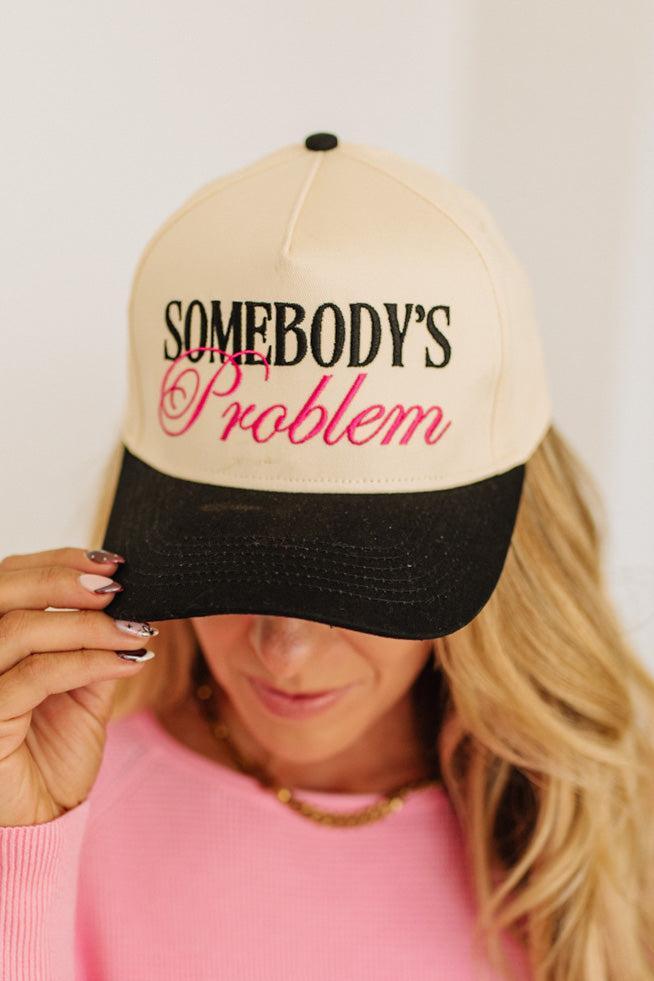 Blonde woman wearing a cream and black trucker hat with "SOMEBODY'S PROBLEM" embroidered at the front in pink and black.