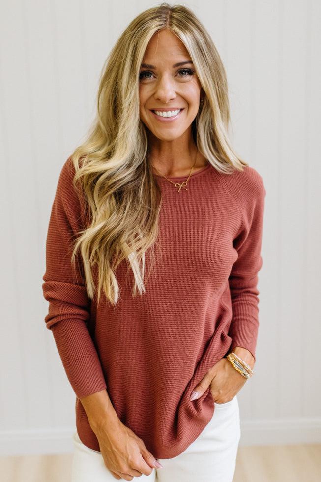 A smiling blonde woman wearing a ribbed long-sleeve crewneck sweater in a dusty terracotta rose color, paired with white pants, standing against a light-colored paneled wall.