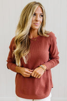 A woman with long blonde hair wearing a ribbed, long-sleeve crewneck sweater in a muted terracotta or rust color, paired with white pants. She is standing against a white paneled wall.