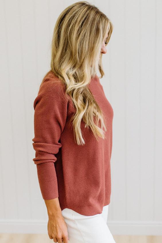 A woman with long blonde hair is shown from a side-back angle wearing a ribbed, dusty rose long-sleeve sweater and white pants against a white paneled wall.
