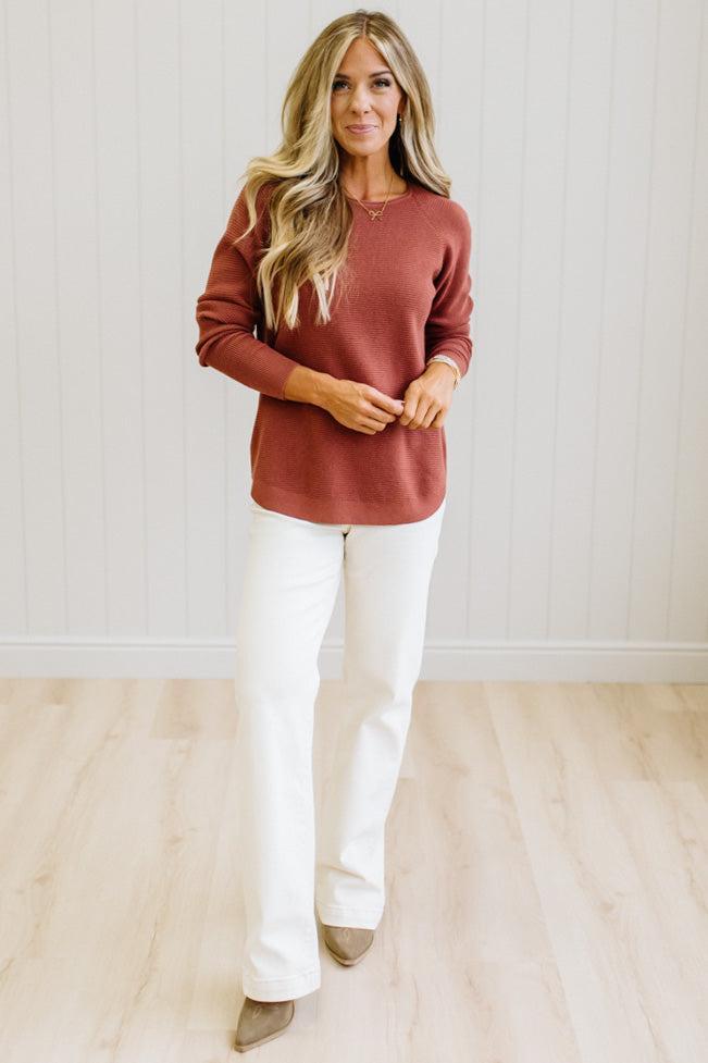 A woman shown from the back wearing a textured, ribbed knit long-sleeve sweater in a dusty rose or terracotta color, paired with white pants against a white paneled wall.