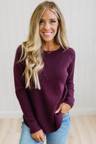 A smiling blonde woman wearing a long-sleeve ribbed knit sweater in deep burgundy and light blue jeans, standing against a white paneled wall.
