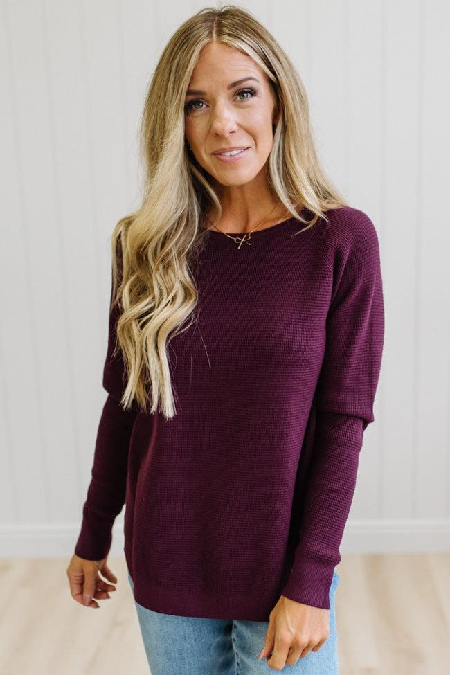A woman with long blonde hair wearing a ribbed long-sleeve crewneck sweater in deep burgundy, paired with blue jeans, standing against a white paneled wall.
