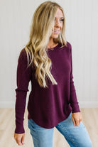 A woman with long blonde hair wearing a ribbed long-sleeve knit top in deep burgundy and light blue jeans, standing in a brightly lit room with white paneled walls and light wood flooring.