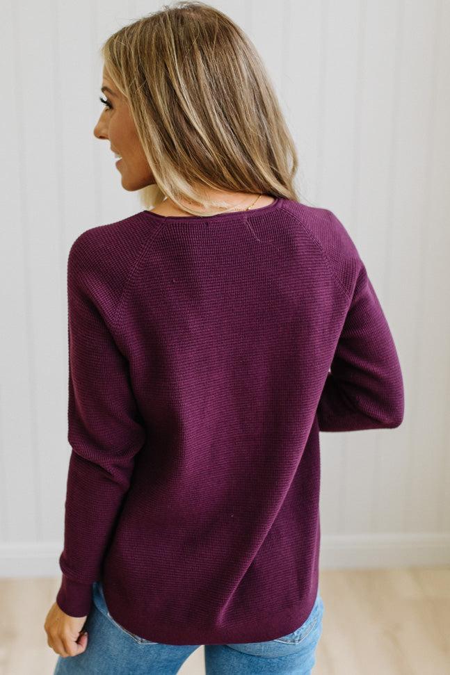 A woman with blonde hair is shown from behind wearing a textured, long-sleeve knit sweater in a deep plum purple color, paired with blue jeans, against a white paneled wall.