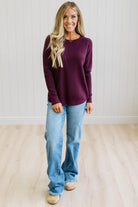 A woman with blonde hair stands in a room with white paneled walls and light wood floors, wearing a long-sleeve crewneck sweater in deep burgundy and light-wash blue jeans.