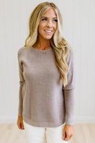 A smiling blonde woman wearing a long-sleeve crewneck knit sweater in a taupe-brown color, paired with white pants, standing against a light-colored paneled wall.